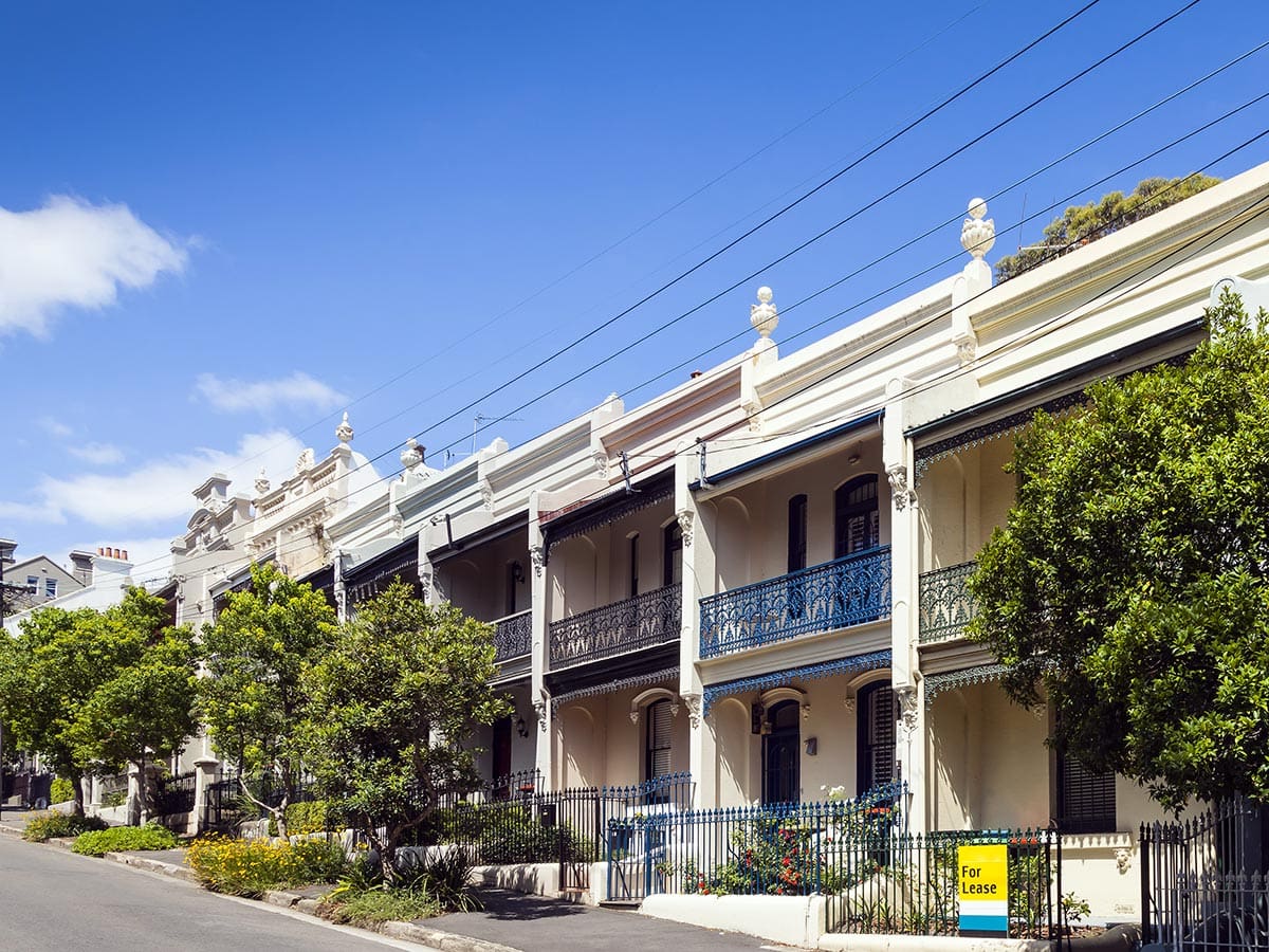 Fila de casas adosadas victorianas con balcones de hierro forjado y letrero de alquiler en una calle soleada de Australia.